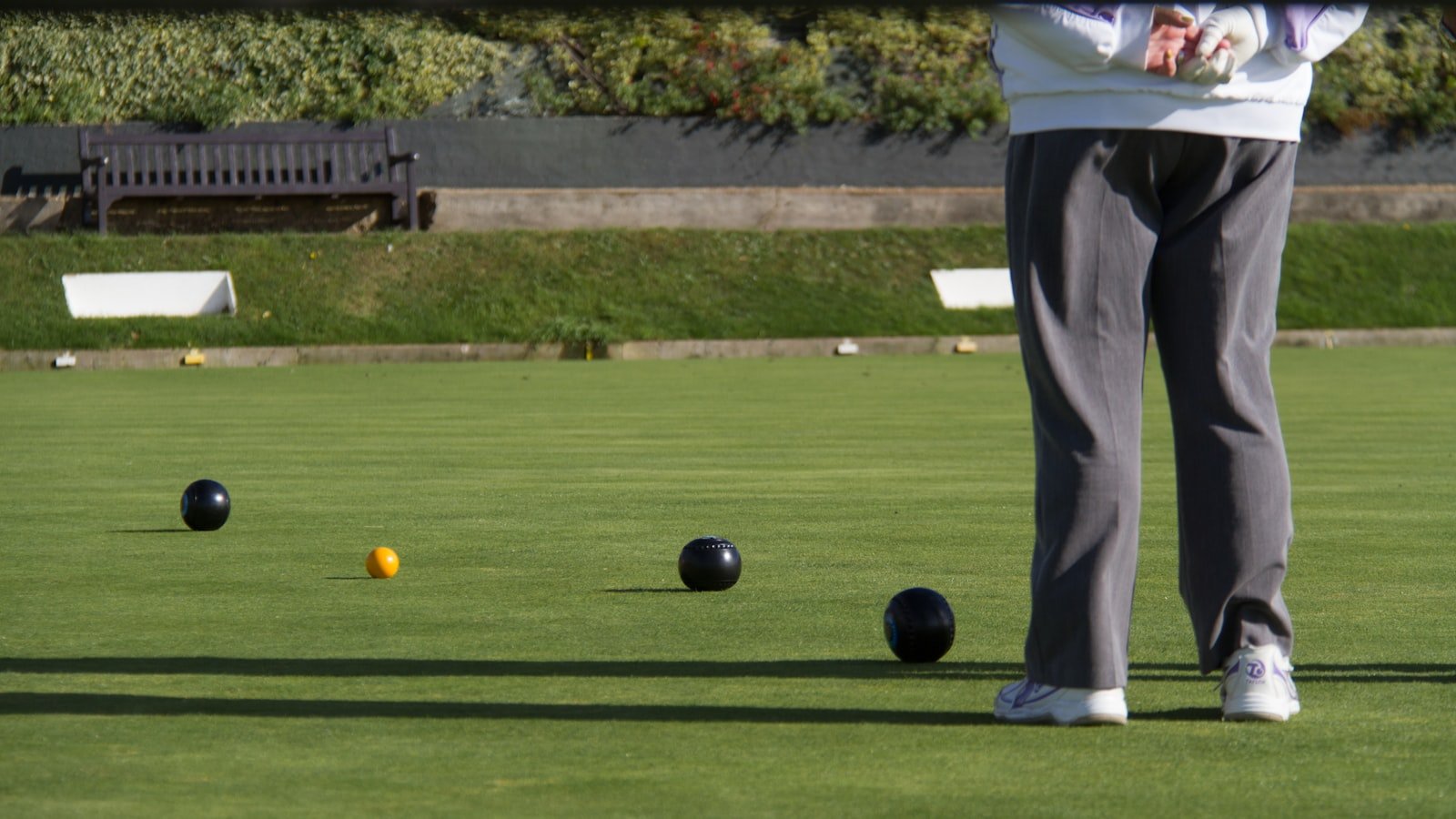 A lawn bowler standing on a manicured green beside a bench, with bowls and the jack in play